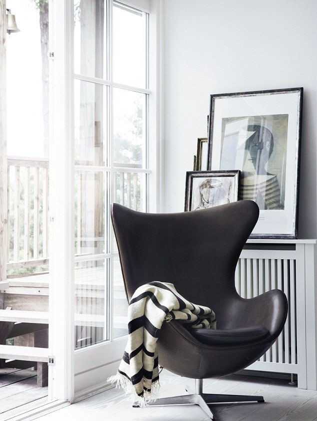 Arne Jacobsen - Chaise Egg en cuir noir sur un pied métal chromé. Il est dans un angle d'une pièce blanche avec des cadres derrières et une baie vitrée ouverte. Une couverture rayée blanche et noire avec des franges est posée sur le fauteil.